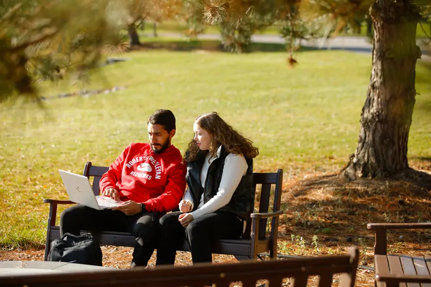 2 students sit on a bench and look at a laptop together